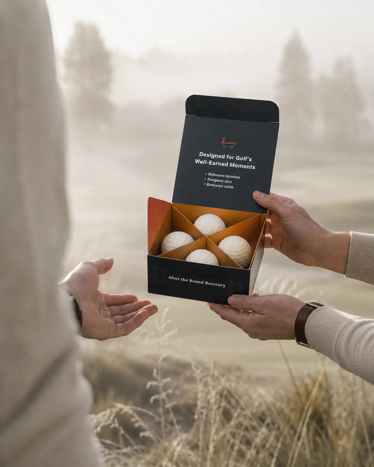A person holding an open black gift box containing four golf balls, with another person's hands assisting in an outdoor field setting with a soft-focus background.
