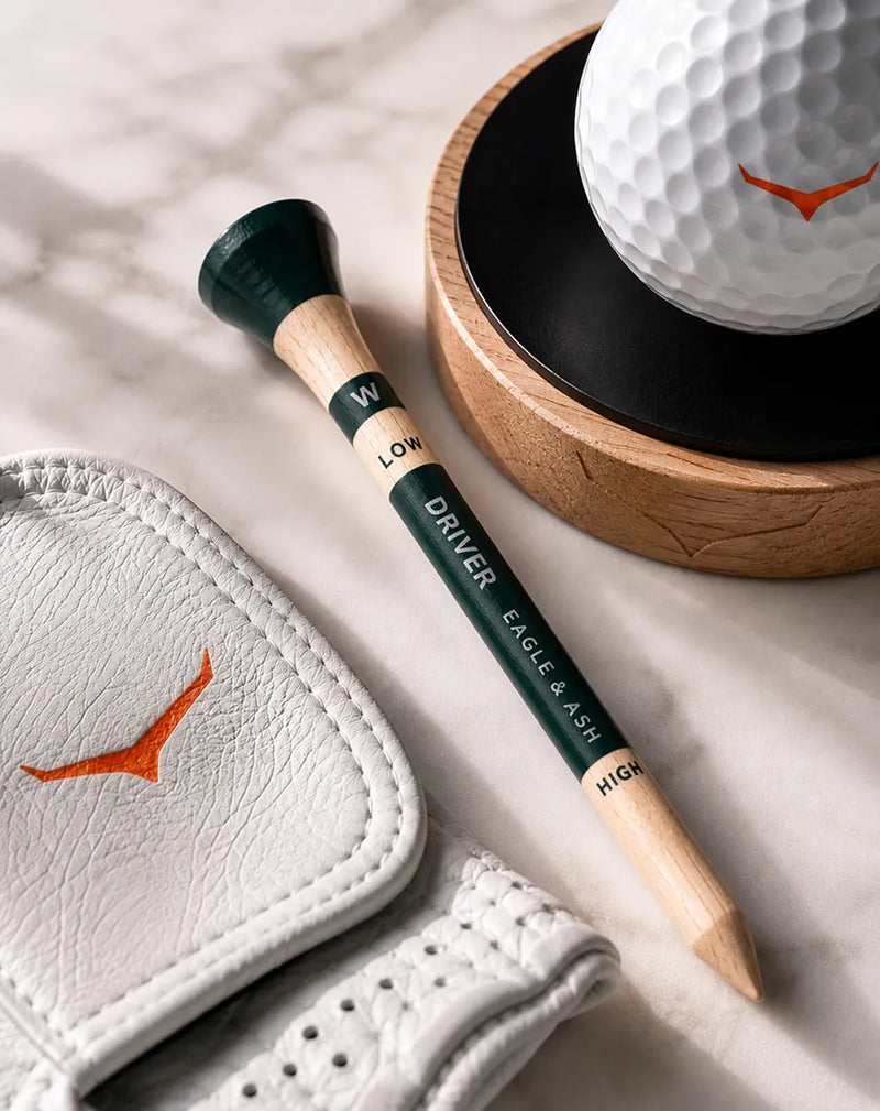 A golf club fitting/alignment tool with a dark green and light wood handle lies on a white leather golf glove beside a white golf ball on a wooden stand, all on a light marble surface.