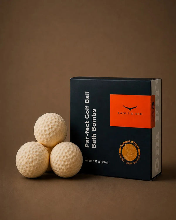 A box of Eagle & Ash Par-fect Golf Ball Bath Bombs next to three golf ball-shaped bath bombs on a solid brown background.