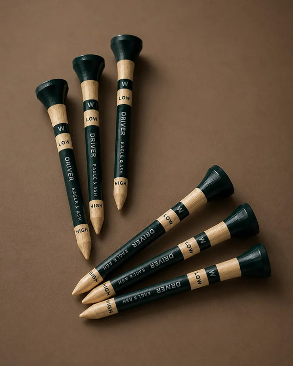 Five golf tees arranged on a smooth brown surface, with three standing upright and two lying diagonally. The tees are dark green with light wood sections and printed text including 'W', 'LOW', 'DRIVER', 'FACE & BACK', and 'HIGH'.