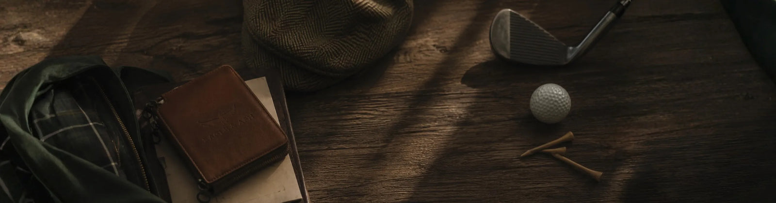 A golf club, golf ball, tees, and a brown leather golf bag or case arranged on a rustic wooden table with warm sunlight and shadows.