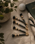Four golf tees with black and gold shafts and white text, arranged on a beige towel alongside a golf ball, a potted plant, and a dark green golf book.