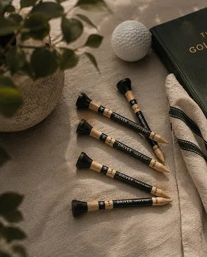 Four golf tees with black and gold shafts and white text, arranged on a beige towel alongside a golf ball, a potted plant, and a dark green golf book.