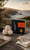A box of Kicker & Co. Par-Fect Golf Ball Bath Bombs sits on a rustic wooden table outdoors, styled with three golf balls, a folded towel, a book, and sunglasses, with a scenic coastal cliff and ocean in the background at sunset.