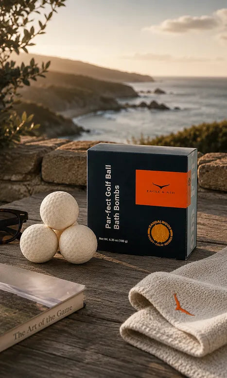 A box of Kicker & Co. Par-Fect Golf Ball Bath Bombs sits on a rustic wooden table outdoors, styled with three golf balls, a folded towel, a book, and sunglasses, with a scenic coastal cliff and ocean in the background at sunset.