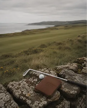 A brown leather golf accessory, likely a wallet or card holder, lies on a stone wall beside a golf club and a golf ball, with a scenic coastal grassy landscape in the background.