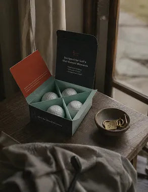 A dark teal and orange gift box containing four bath bombs, styled on a rustic wooden table next to a small bowl of gold keys.