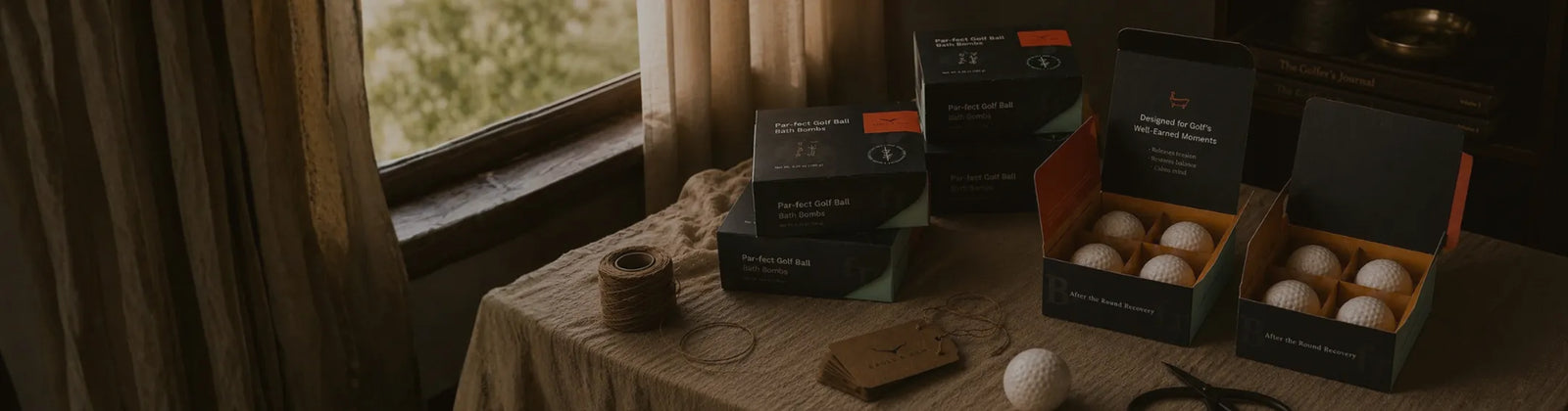 Three open black gift boxes containing white golf balls are arranged on a rustic tablecloth next to a window in a dimly lit room. A golf club head and a single golf ball rest on the table, with a small wooden tag nearby.